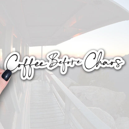 a hand holding a sticker with the text Coffee Before Chaos written on it in black font with white outline.