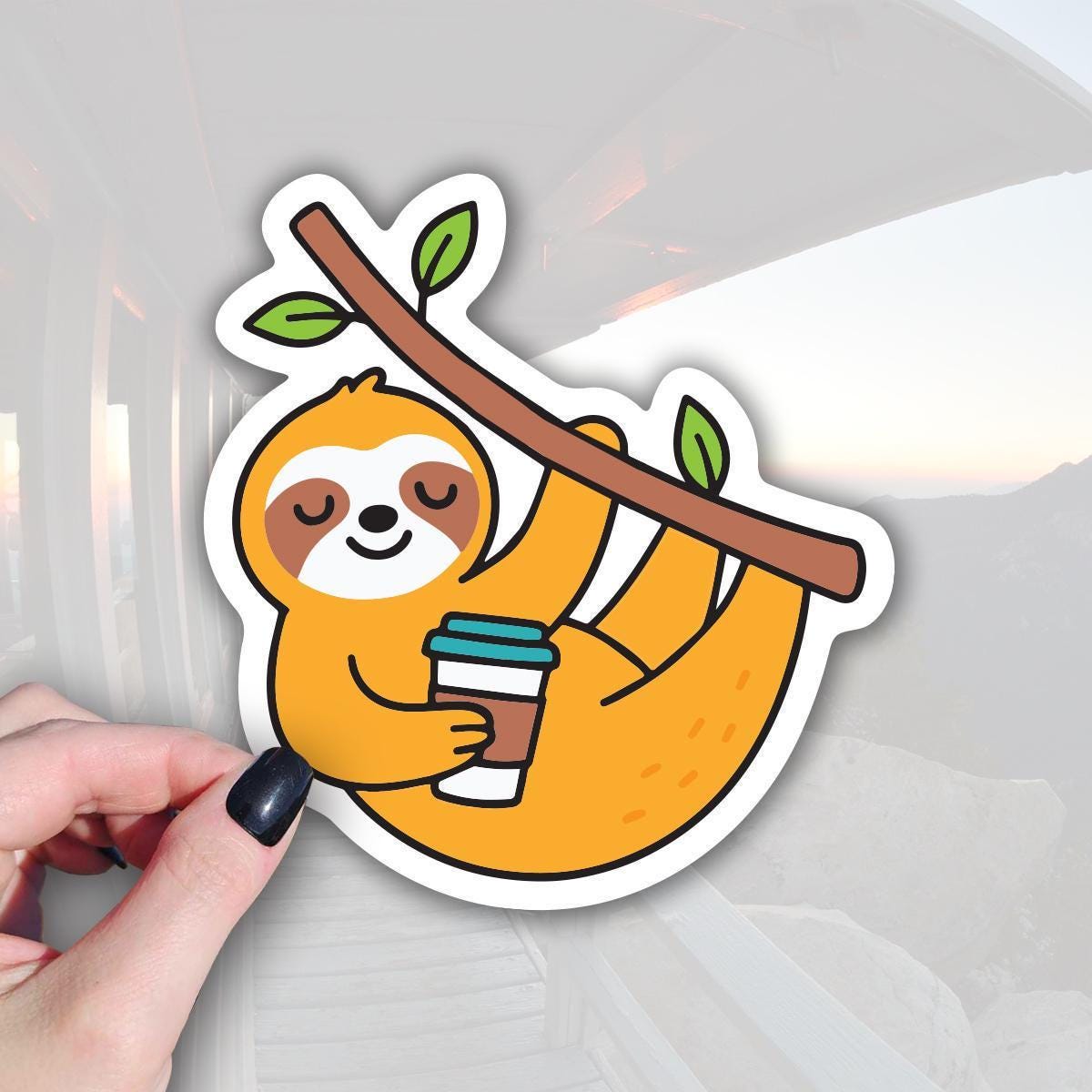 A hand holding a sticker depicting a sloth hanging from a branch with a cup of coffee.