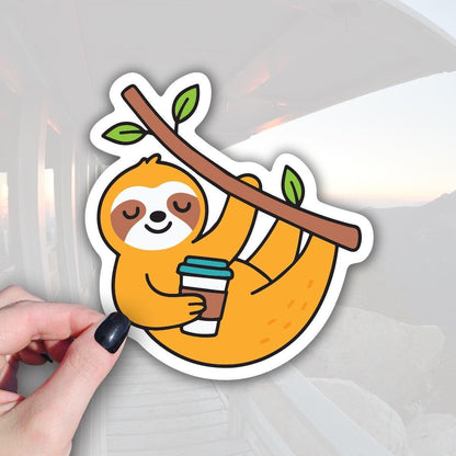 A hand holding a sticker depicting a sloth hanging from a branch with a cup of coffee.